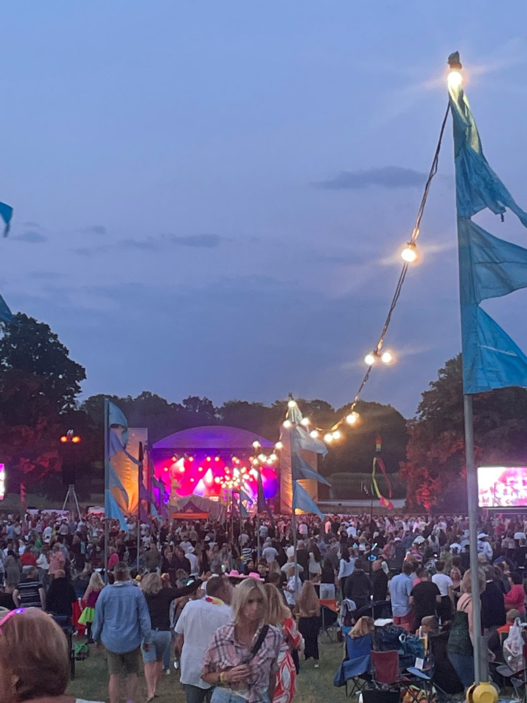 Outdoor stage lit up in magenta with crowds of people, flags and festoon lights for Ibiza Classics concert at Blickling Hall