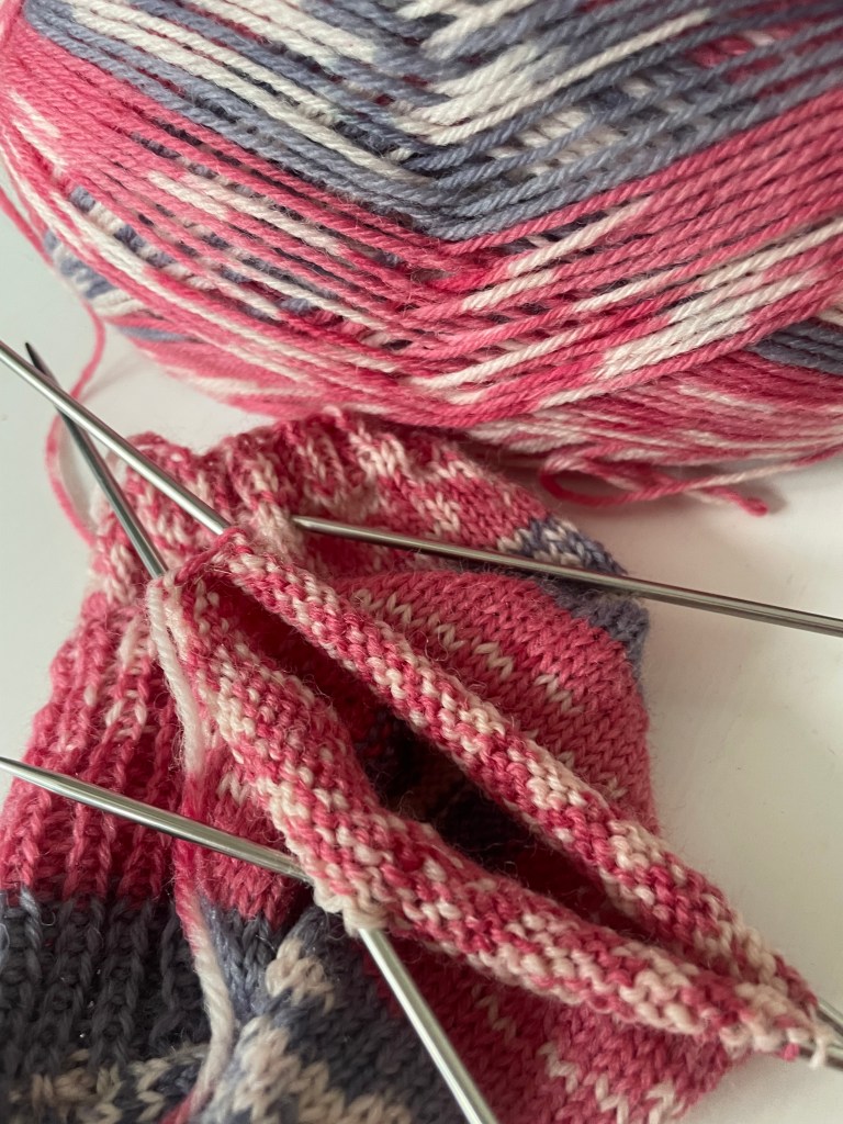 a ball of pink and grey wool beside a part-knitted sock on dpn's